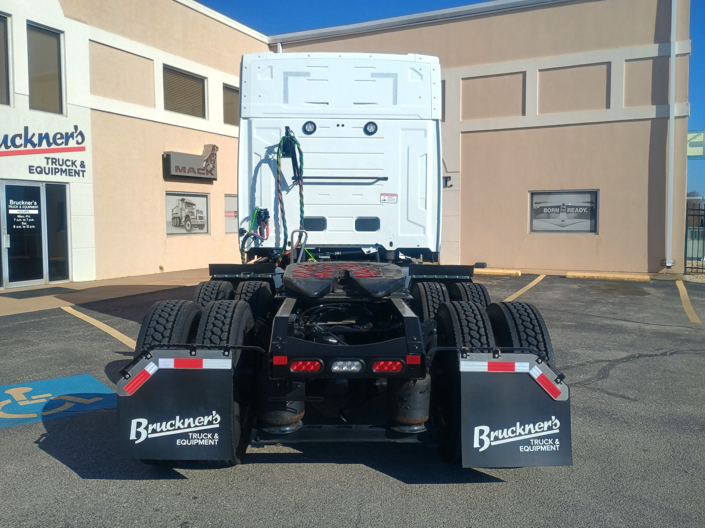 Truck rear view of a white 2027 Mack AN64T64 tractor in a parking lot, showing dual rear axles, mud flaps with Bruckner’s Truck & Equipment logos, and safety lights.