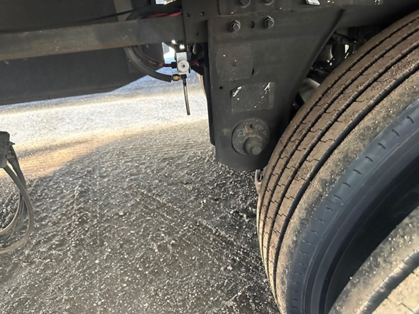 Suspension area of a trailer chassis showing a leaf spring mount and wheel with dual tires; some wiring and metal brackets visible.