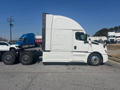 Tractor truck side view showing white Freightliner Cascadia 126 sleeper cab with chrome fuel tanks and blue truck in background.