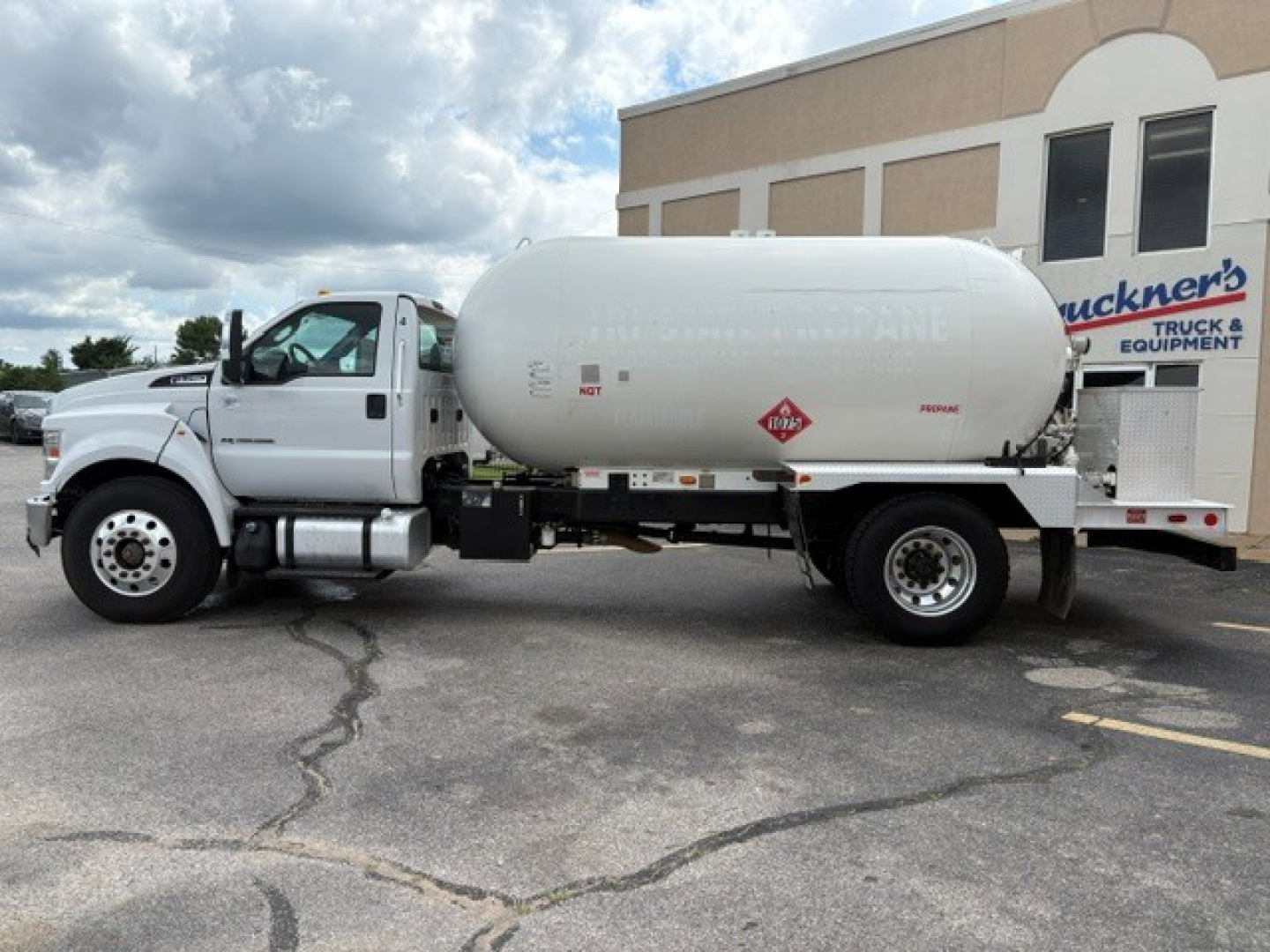 2016 Ford F-750 vacuum/propane or liquid tank truck, white cab, large cylindrical tank mounted behind cab, with hazard placards and small stickers on tank, dealership building in background.