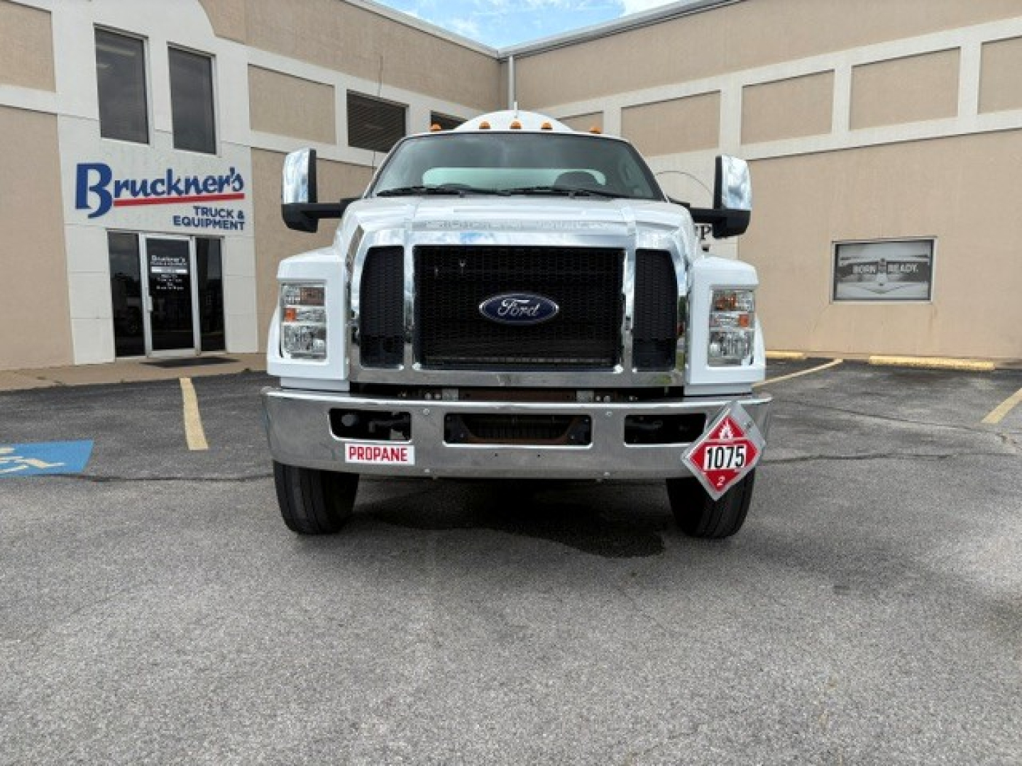 Ford F-750 front view showing large chrome grille, bumper, and dual side mirrors; hazard placard and propane sign on bumper; dealership building in background.