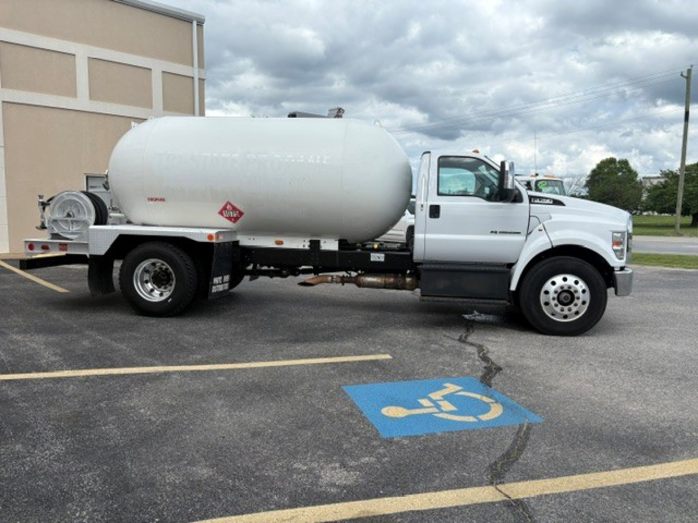 Ford F-750 utility truck with a large cylindrical tank mounted on the chassis, white cab and tank with hazard placards, in a parking lot.