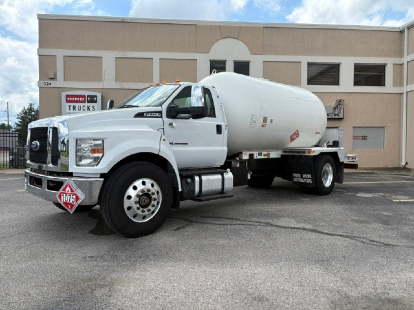 Ford F-750 truck with a cylindrical tank in front of a beige building, front left view showing Hazmat placard and fuel tank steps.