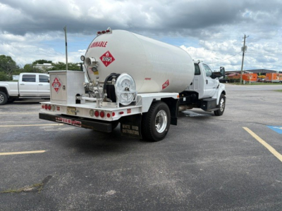 Fuel or propane tanker truck rear view with cylindrical tank and hazard diamonds, U.S. truck chassis, license plate area, and company equipment in lot.