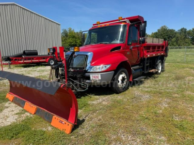 Red 2007 International 4300 plow truck with large front plow, dump bed, and amber warning lights, parked on grass near a metal building.