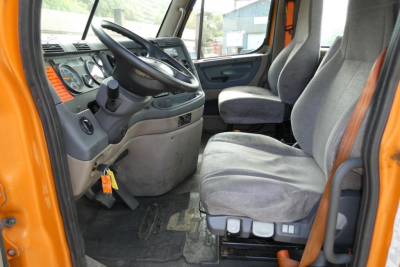 Dashboard and driver seat area of a Freightliner Cascadia cab with gray seats, orange exterior, steering wheel, pedals, and instrument cluster. Visible signs include a large steering wheel and control panels.