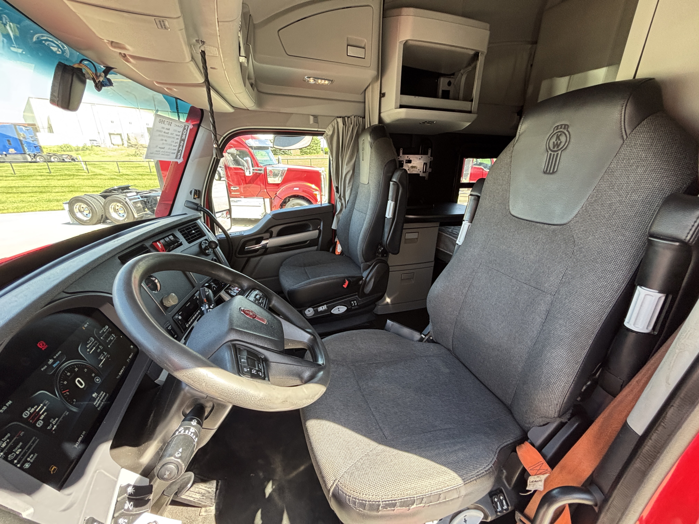 Truck cab interior of a Kenworth T680 showing steering wheel, driver's seat with Kenworth logo, dashboard gauges, and a sleeper area behind.