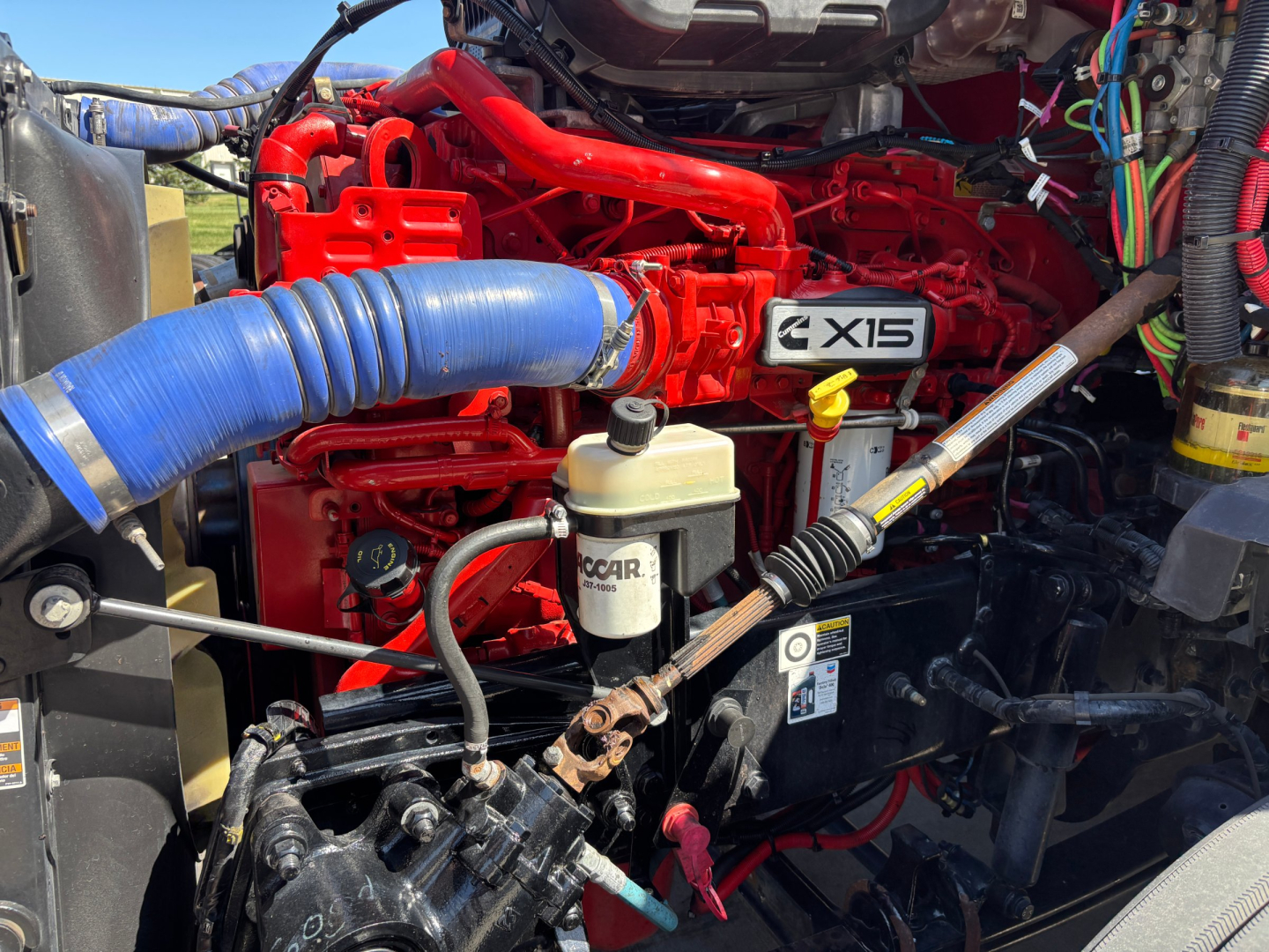 Kenworth T680 engine bay showing X15 badge, red engine, blue intercooler hose, brake reservoir, and wiring harnesses.