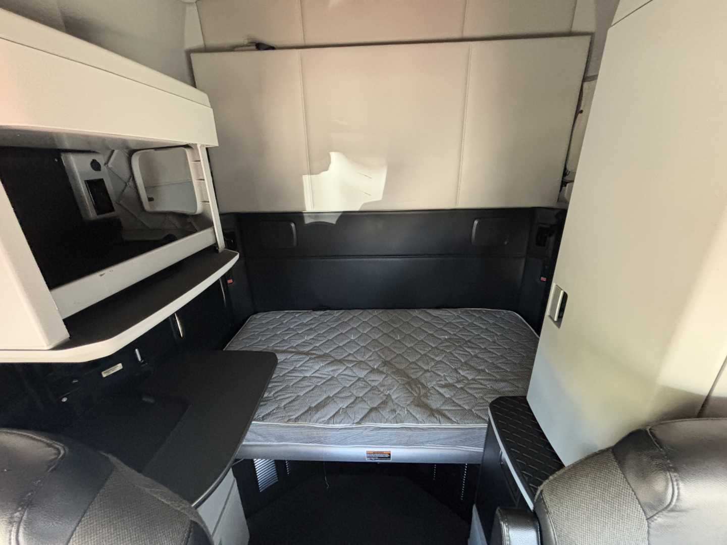 Kenworth T680 sleeper cab interior showing bunk bed mattress with under-bed storage and side cabinet; upper bunk area and doorway visible.