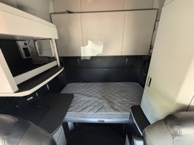 Kenworth T680 sleeper cab interior showing bunk bed mattress with under-bed storage and side cabinet; upper bunk area and doorway visible.