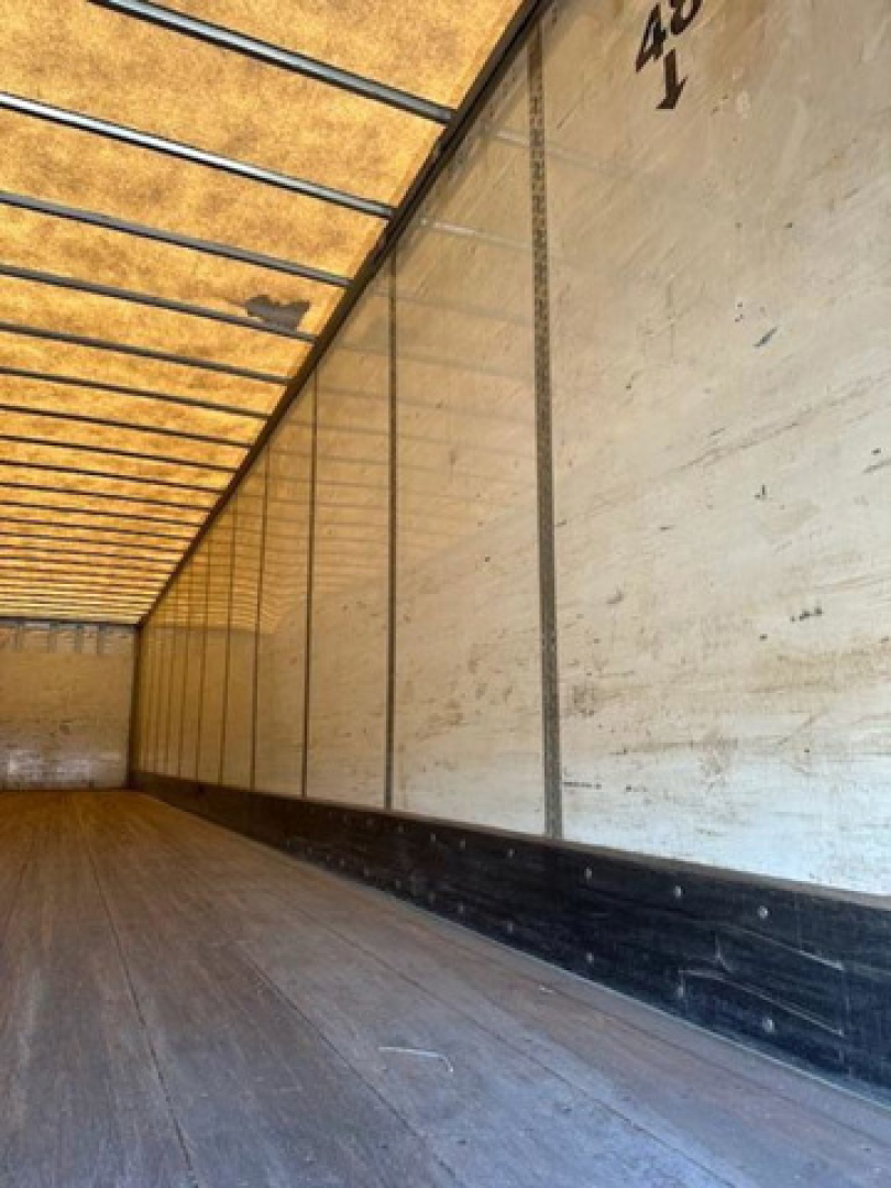 Trailer interior showing wooden floor and sidewall panels with metal rails; long van cargo area, light from open rear door.