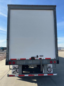 Rear view of a white Wabash van trailer with metal frame, red/white reflective striping, and bumper lights.