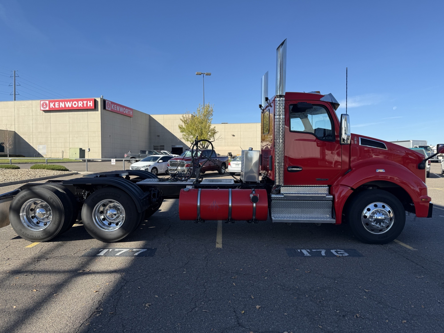 Kenworth T880 conventional day cab truck in red, side profile, with chassis and fuel tanks visible; Kenworth dealership in background.