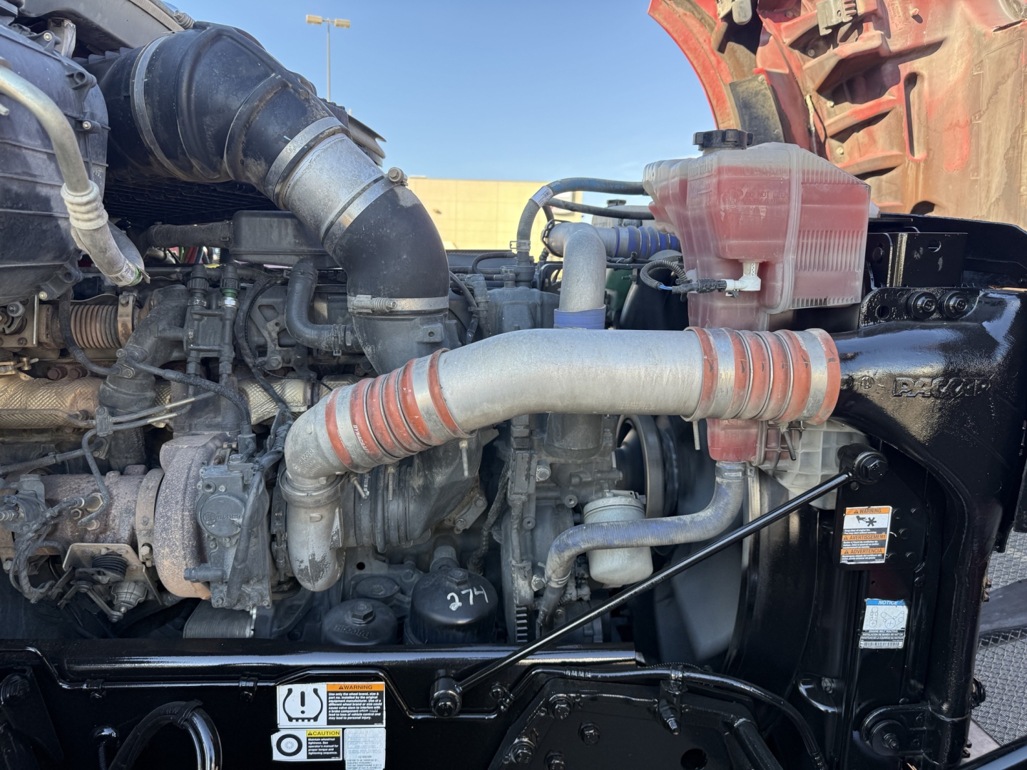 Kenworth T880 engine bay showing air intake hose, turbocharger, coolant reservoir, and various hoses in a daytime truck engine compartment.