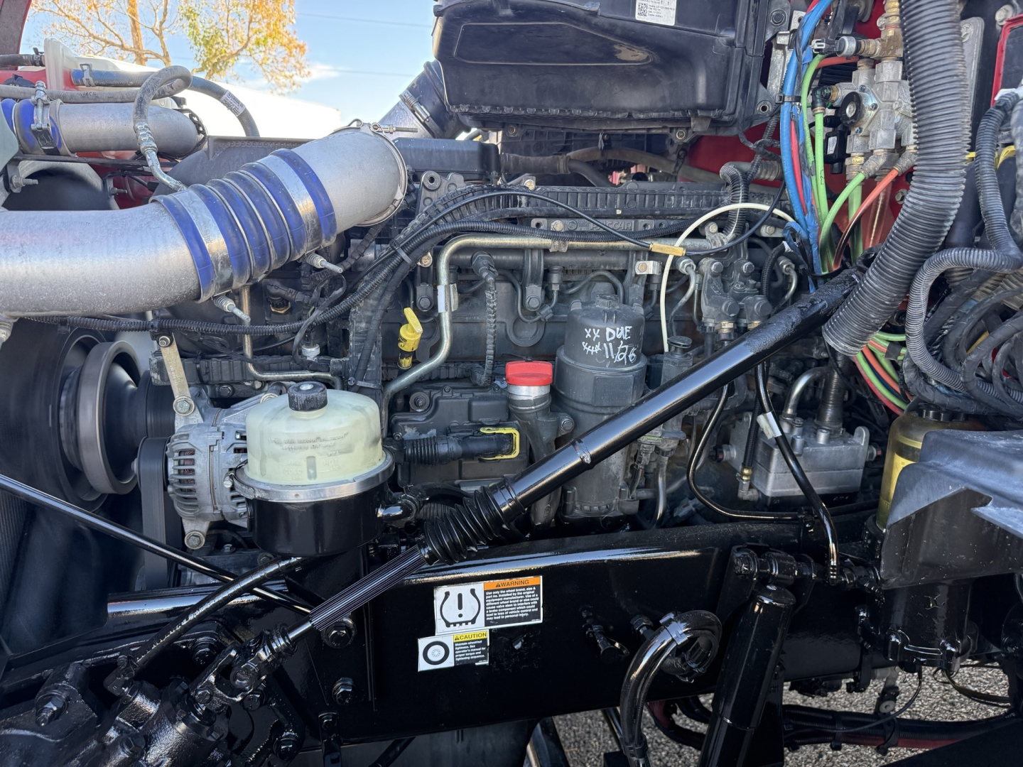 Kenworth T880 engine bay with visible hoses, wiring, and components. Engine block with labeled caps and connectors, battery area, and surrounding frame.