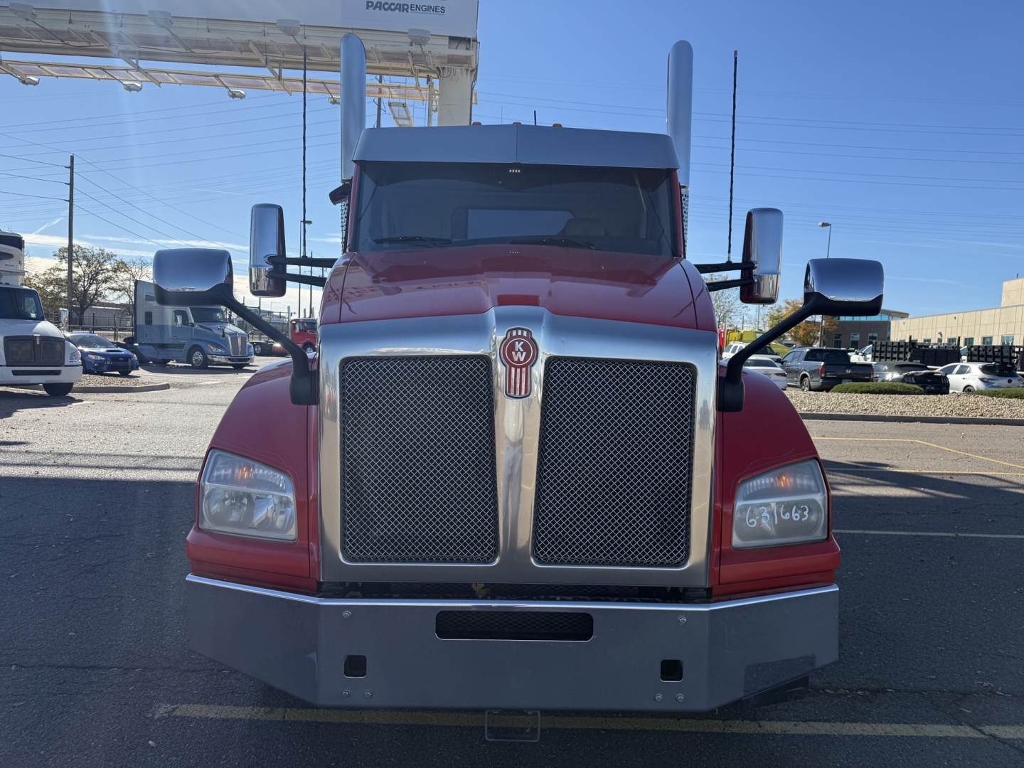 Front view of a red Kenworth truck with large chrome grille, headlights, and bumper. Emblem centered on grille. Side mirrors visible. Clear sky.