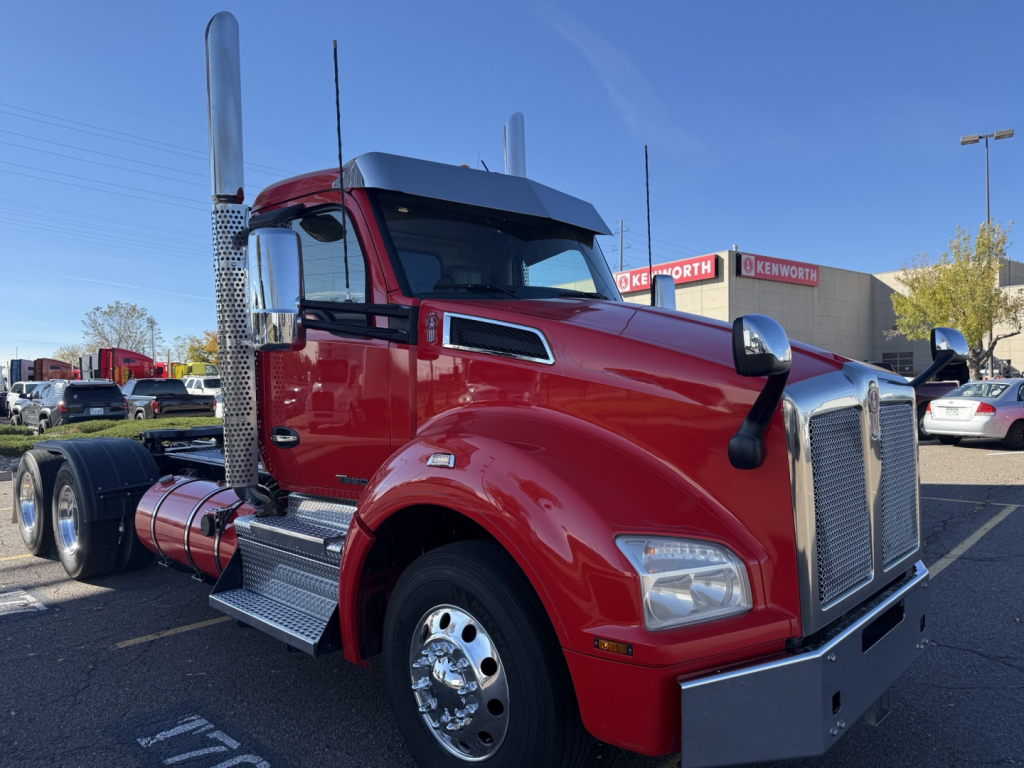 Kenworth T880 conventional daycab truck, red exterior, chrome exhaust stacks, large chrome grille, silver bumper, side steps, dual rear axles visible, dealership lot in background.