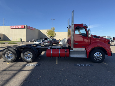 Kenworth T880 conventional day cab truck in red, side profile, with chassis and fuel tanks visible; Kenworth dealership in background.