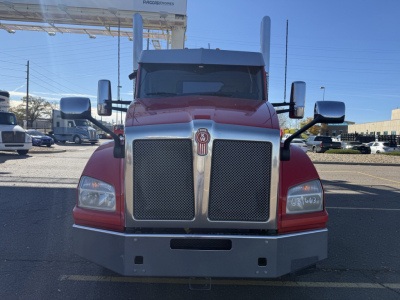Front view of a red Kenworth truck with large chrome grille, headlights, and bumper. Emblem centered on grille. Side mirrors visible. Clear sky.