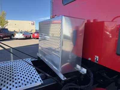 Diamond plate toolbox mounted on the side of a red Kenworth truck with a metal storage box and access steps in the foreground.