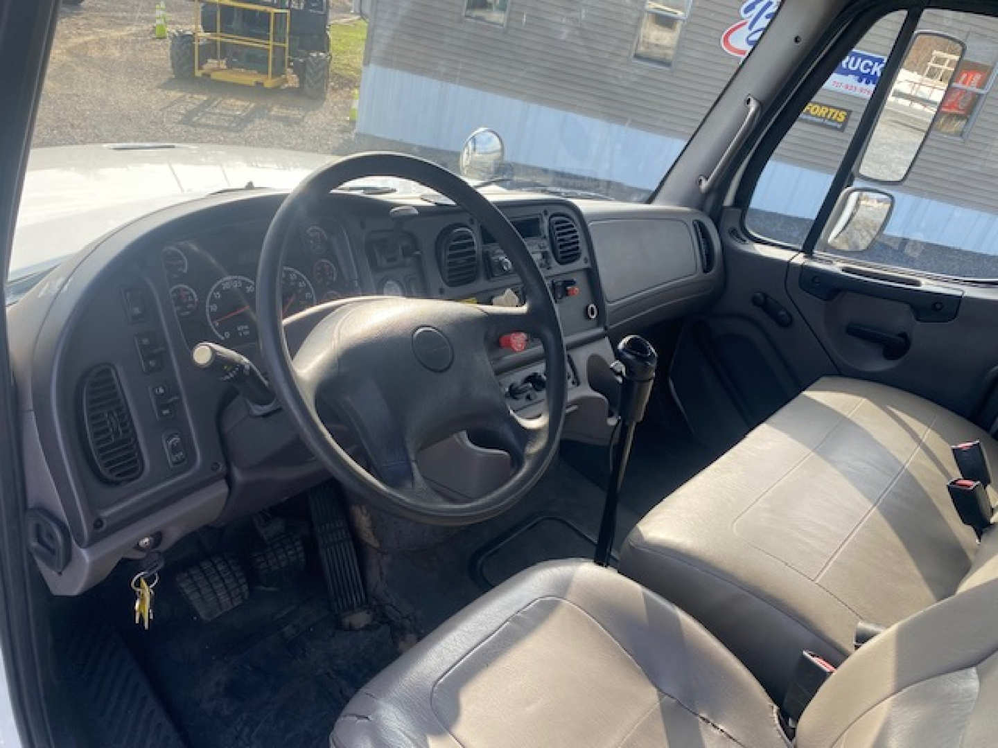 Truck cab interior of 2011 Freightliner M2-112 showing steering wheel, dashboard gauges, manual gear shifter, and beige seats.