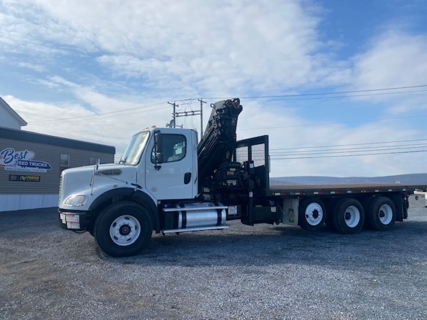 Truck cab and long-bed crane truck side view showing Freightliner M2-112 with crane mounted, white cab, three-axle rear, dealership lot.