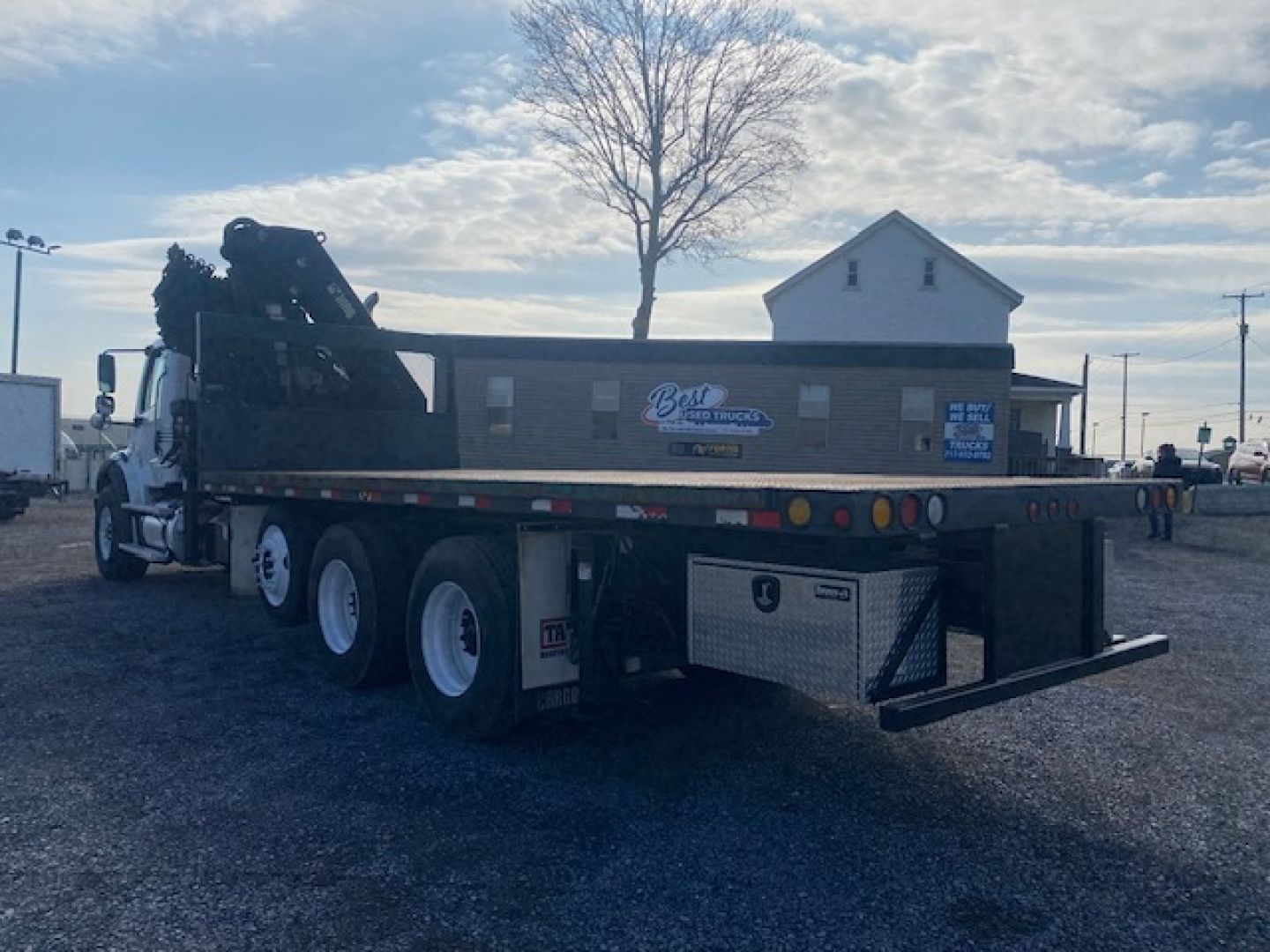 Truck rear-left side view of a Freightliner M2-112 flatbed crane truck with a crane arm folded on the cab-back. Side decals and a square toolbox visible along the bed.