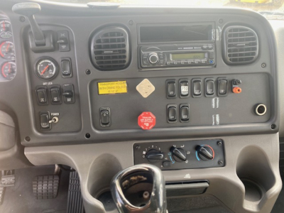 Truck cab dashboard showing steering column, gauges, air vents, radio, and control switches. Visible labels include a yellow warning label, a red knob, and various gray switches.