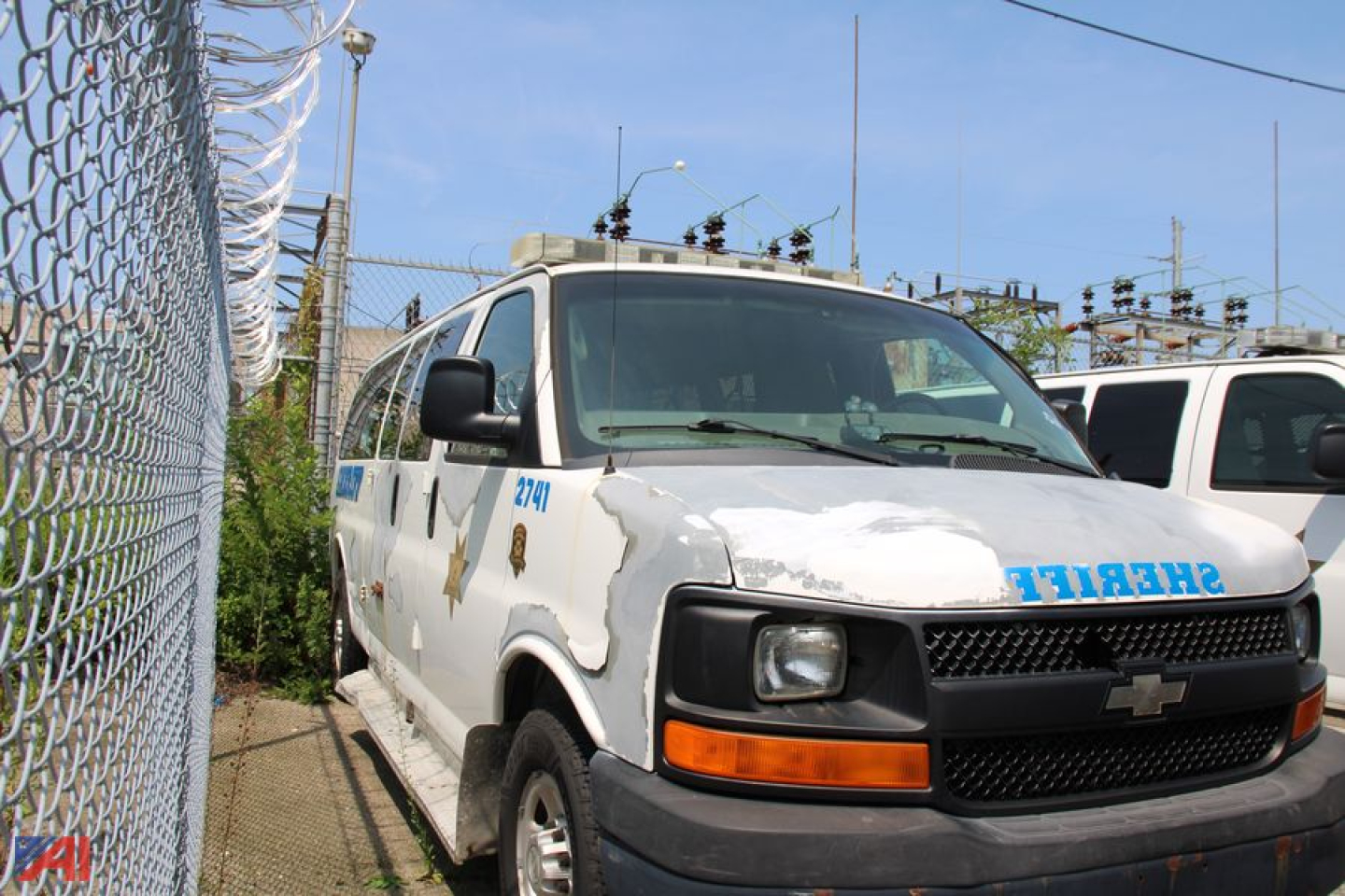 Chevrolet Express van showing damaged hood and front grille with headlights; blue lettering and numbers on side; chain-link fence in background.