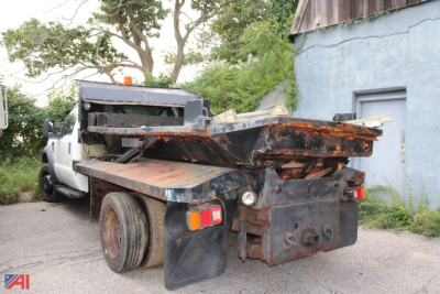 Flatbed tow truck rear view showing rusted metal flatbed and dual rear lights; Ford F-550 truck in background; VIN plate not visible.