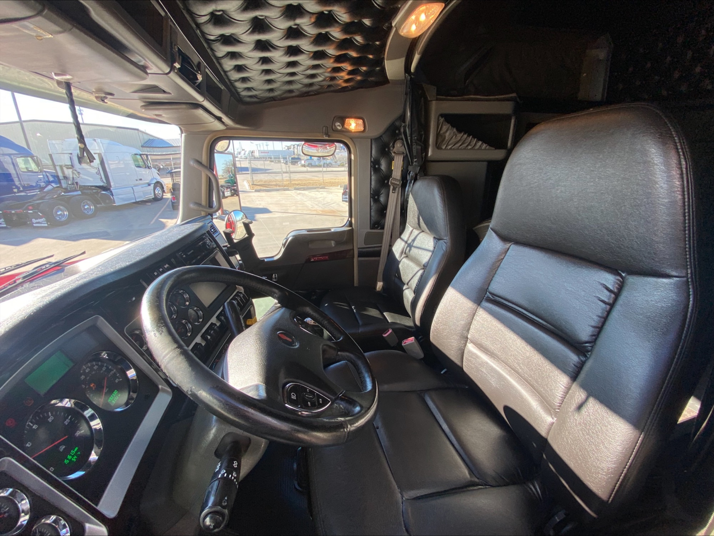 Kenworth W900L truck cabin interior view with steering wheel, dashboard gauges, and driver's seat; window and exterior truck cab visible.