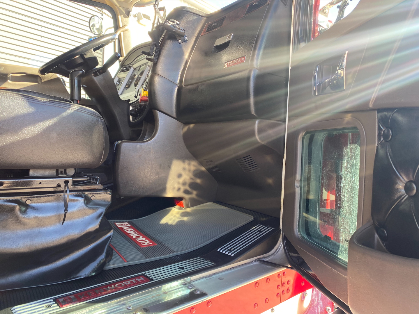 Kenworth W900L cab interior showing driver's seat, steering wheel, dashboard, floor mats, and door sill with Kenworth branding.