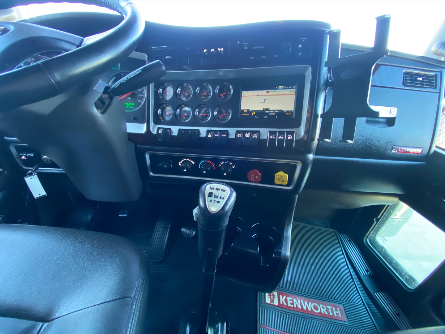 Kenworth W900L truck interior dashboard and gear shifter showing steering wheel, instrument cluster, GPS display, control switches, and Kenworth-branded floor mat.