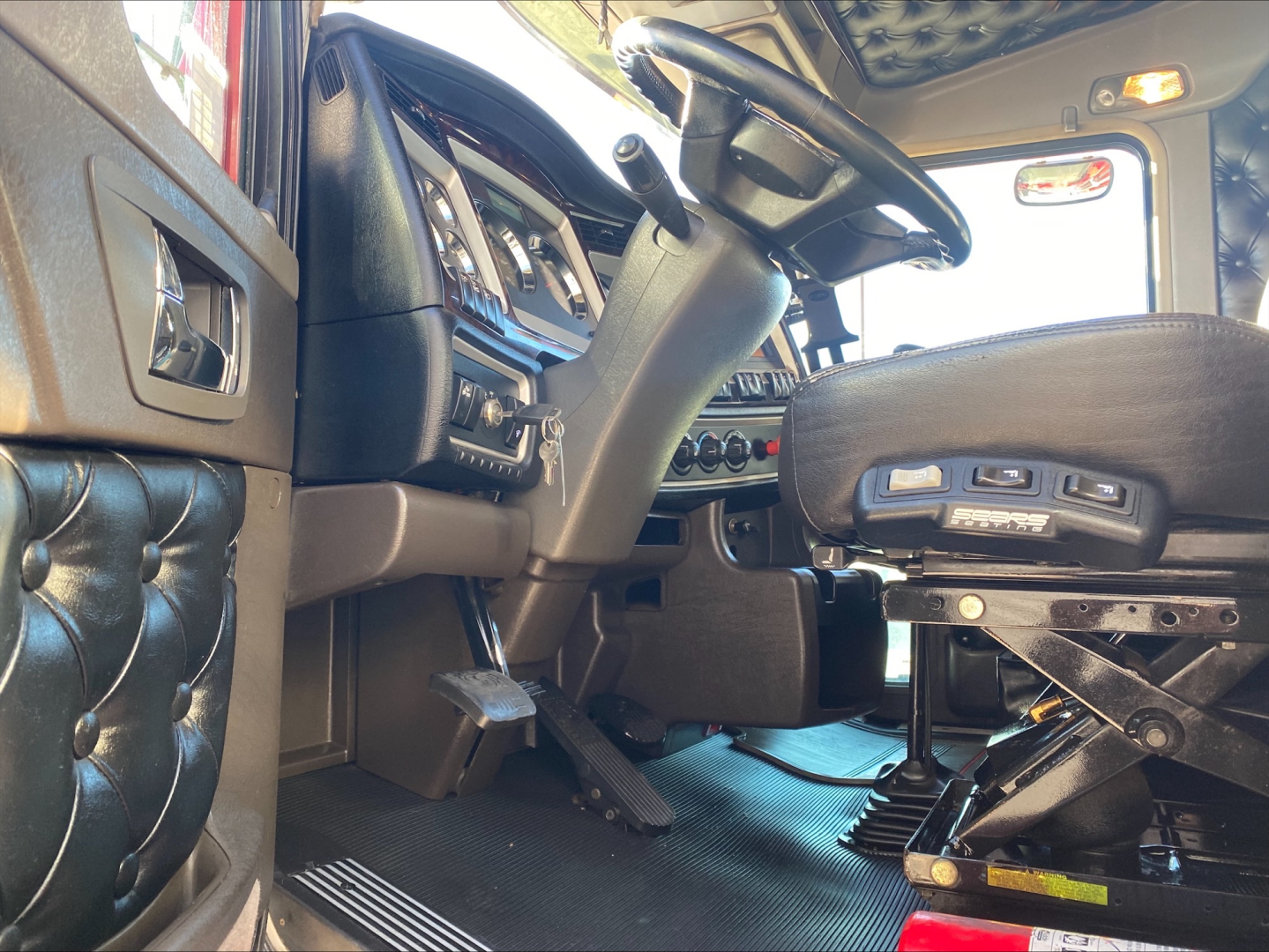 Interior of a Kenworth W900L truck driver area showing steering wheel, dash, and adjustable seat with controls.