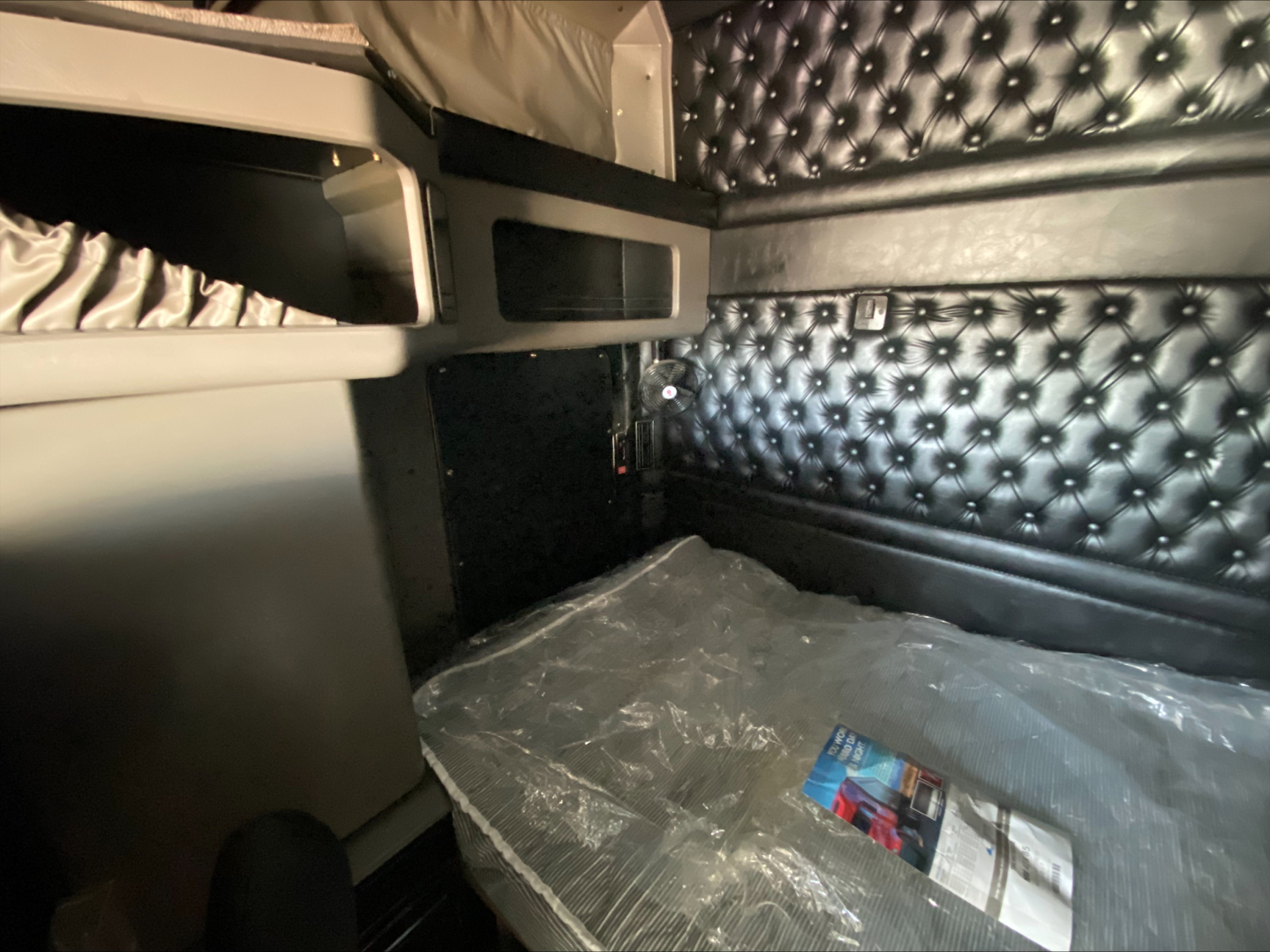 Kenworth W900L sleeper cab interior showing storage compartments and quilted black upholstery with plastics and a small brochure on the floor.