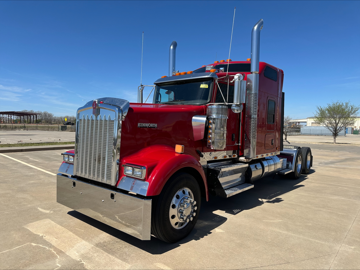 Kenworth W900L conventional sleeper truck in red, facing left, chrome grille, dual stacks, large front bumper, empty lot.