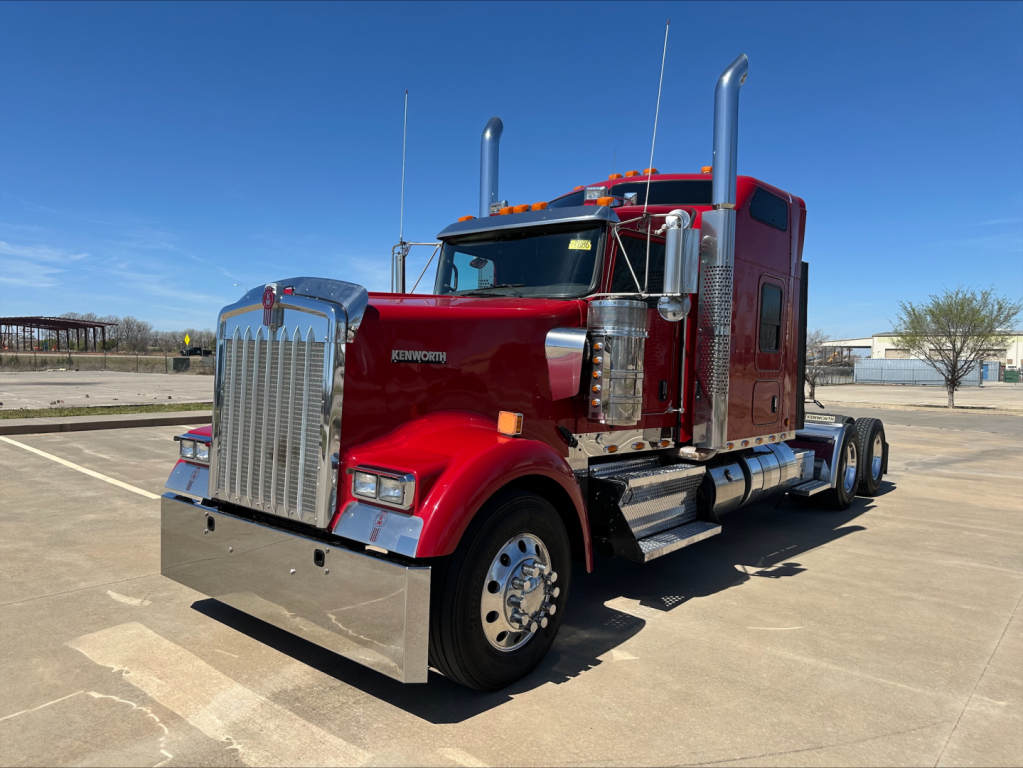 Kenworth W900L conventional sleeper truck in red, facing left, chrome grille, dual stacks, large front bumper, empty lot.