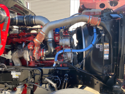 Kenworth W900L engine bay showing large intake/exhaust piping, red engine block, blue hoses, and radiator with fan shroud.