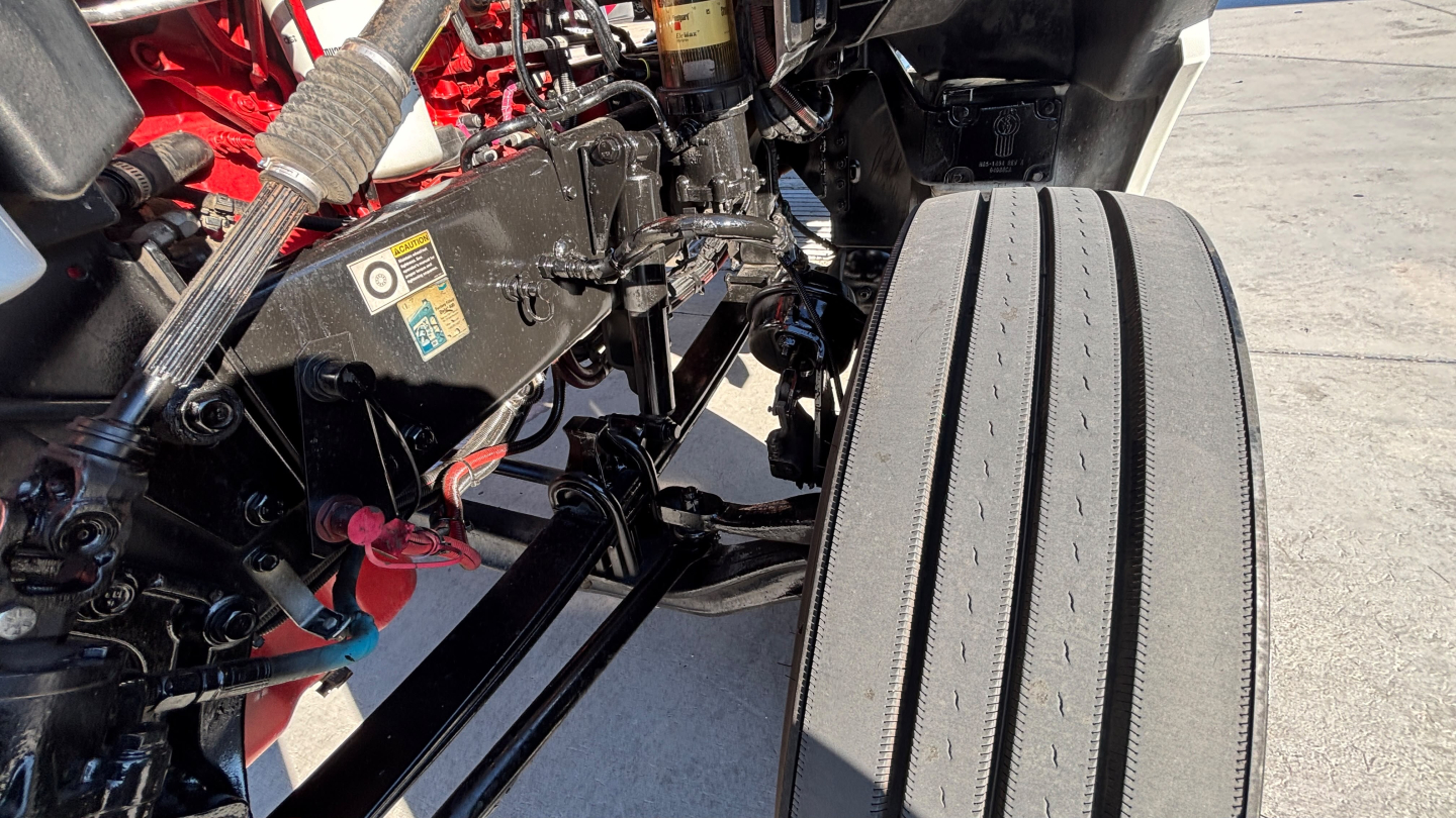 Kenworth T880 chassis and front wheel well showing hydraulic lines and frame; visible components include a black metal frame, red-painted engine block in background, and a tire with tread.