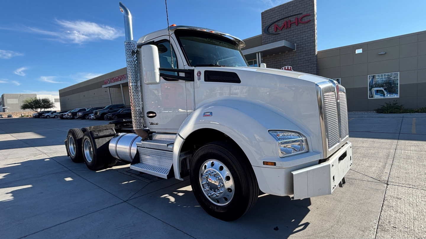 Kenworth T880 conventional daycab truck in a light color, front three-quarter view, large chrome exhaust stack, dual rear axles, and fuel tanks visible.