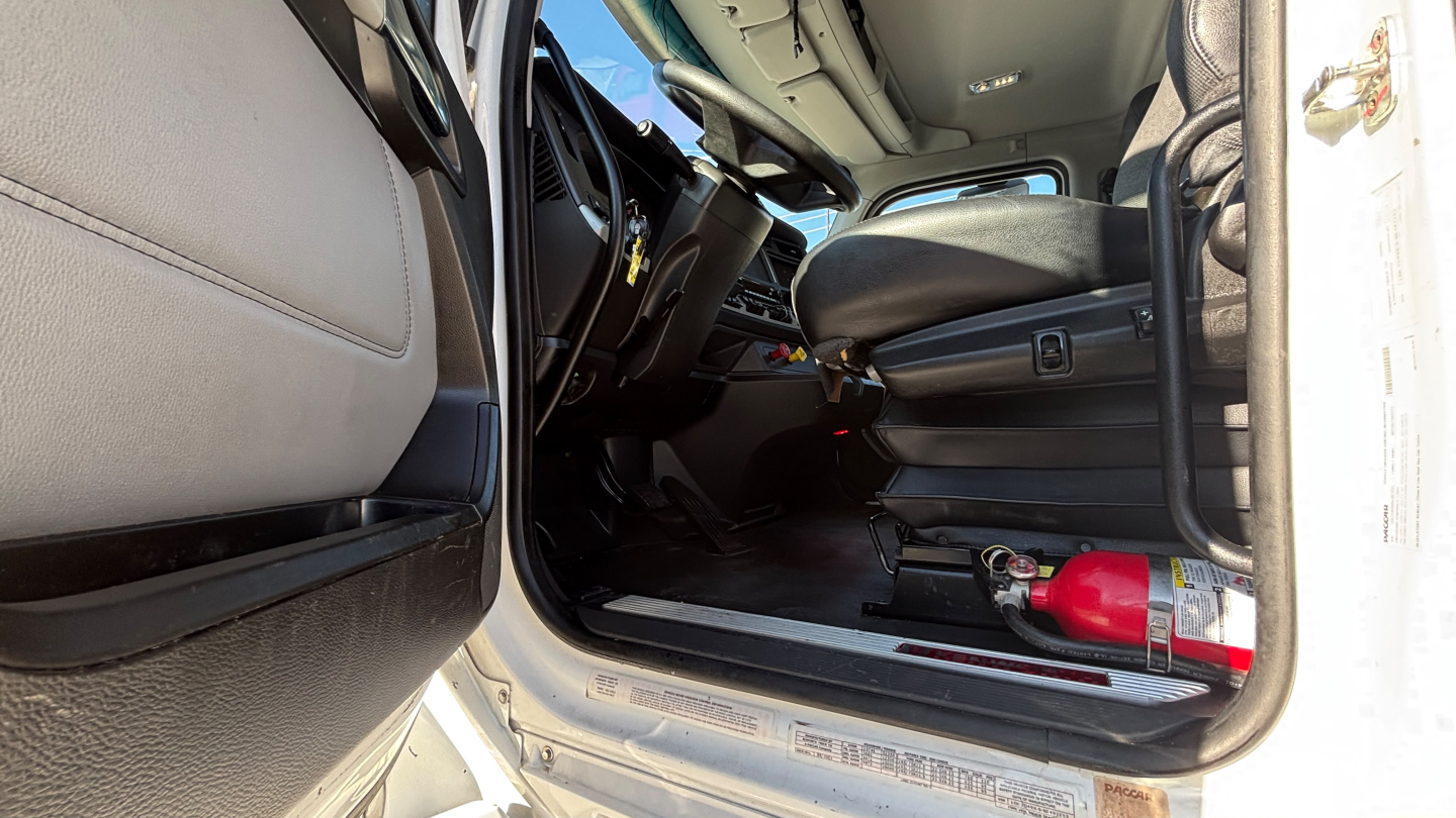 Truck cab interior view of a 2022 Kenworth T880, open driver door, steering wheel, dashboard, seat, and a red fire extinguisher on the floor near the entry.