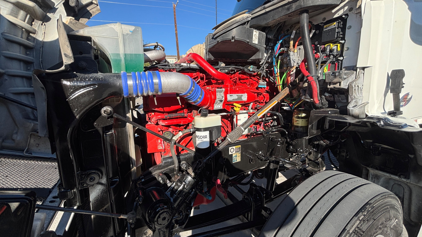 Kenworth T880 engine bay area with red engine block, blue silicone hose, white reservoir, and exposed wiring; front cab open showing electrical and fluid components.