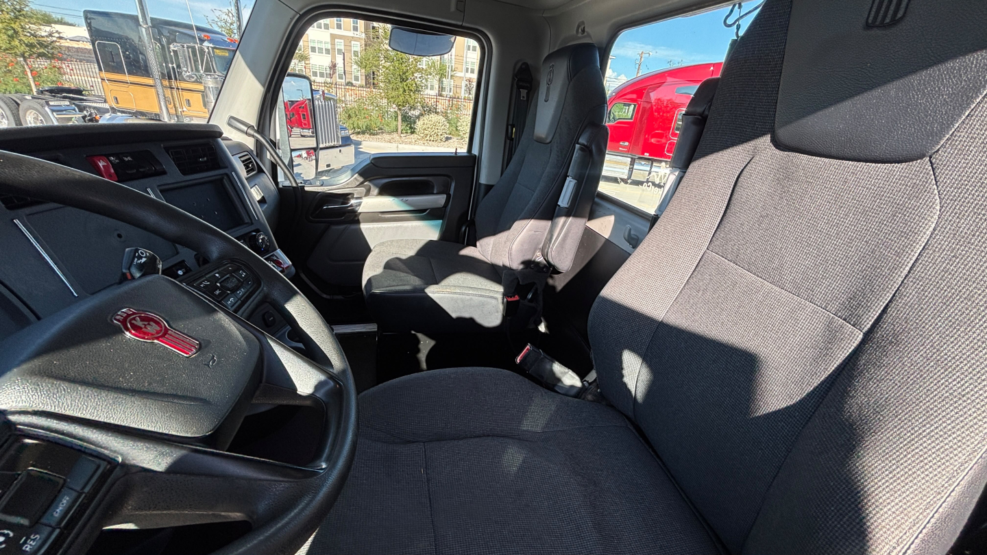 Kenworth T880 truck interior showing grey cloth seats, steering wheel, dashboard, and driver door, front cab with partial passenger seat visible.