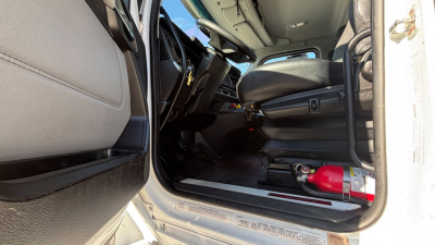 Truck cab interior view of a 2022 Kenworth T880, open driver door, steering wheel, dashboard, seat, and a red fire extinguisher on the floor near the entry.