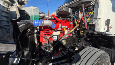 Kenworth T880 engine bay area with red engine block, blue silicone hose, white reservoir, and exposed wiring; front cab open showing electrical and fluid components.