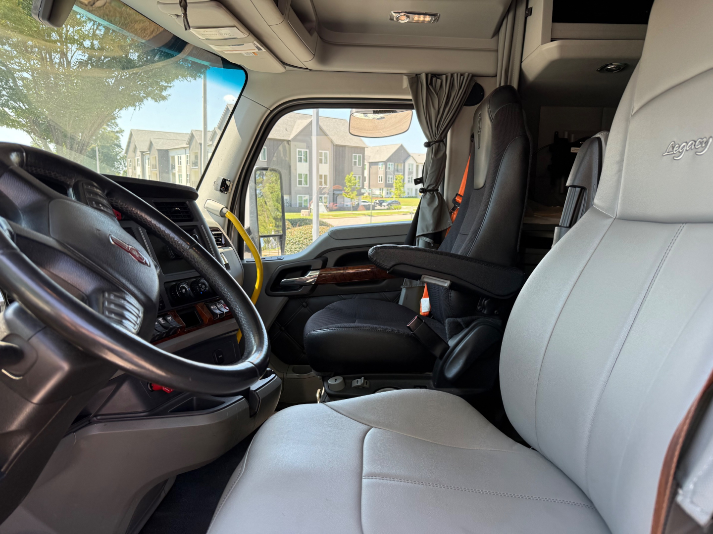 Interior driver area of a 2021 Kenworth T680 with light gray seats, black dashboard, and wooden trim. View from passenger seat showing steering wheel, front seats, and dashboard controls.
