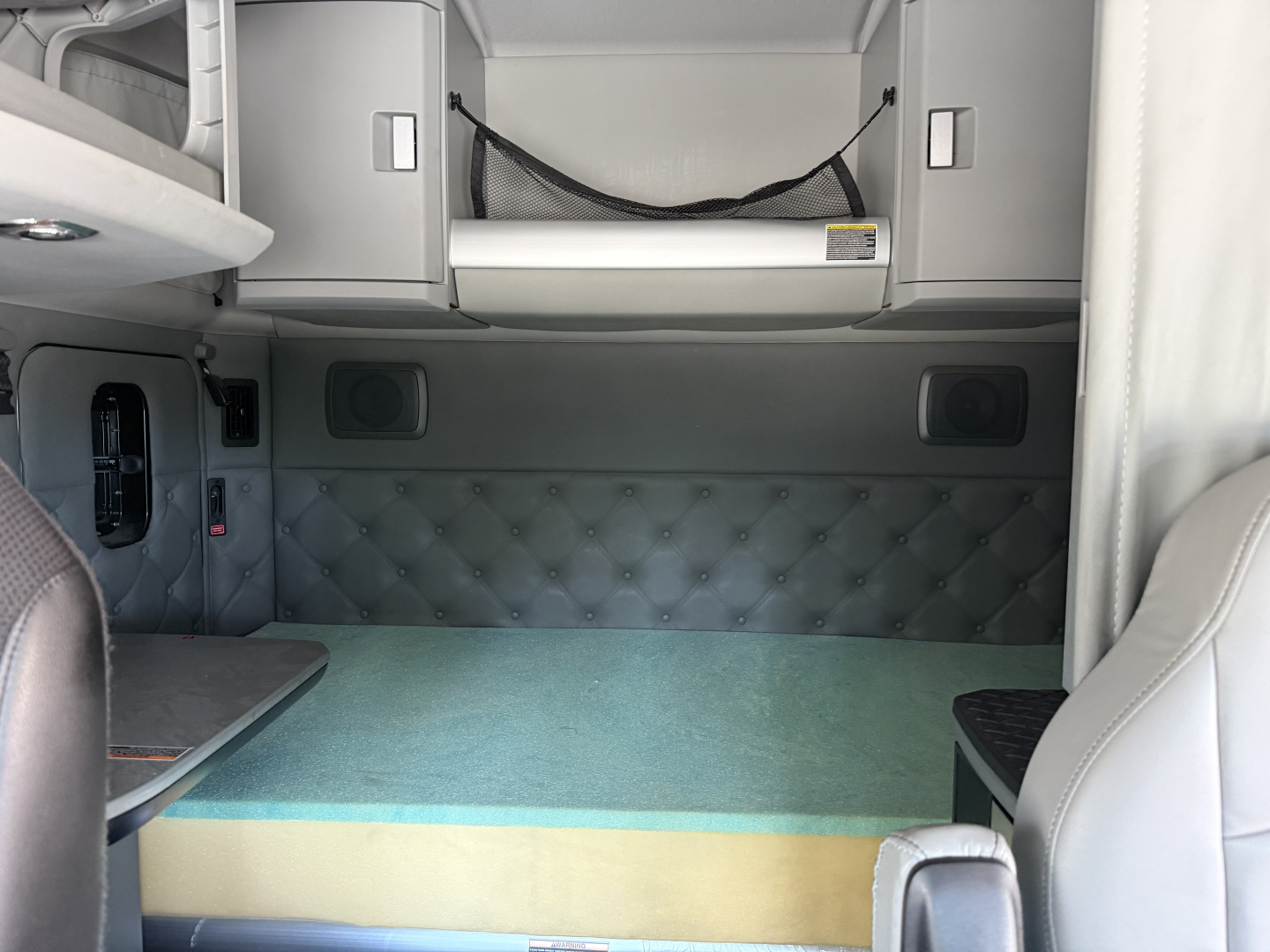 Kenworth T680 sleeper cab interior view with storage compartments and a quilted padded wall; cargo area visible with teal and yellow steps and a netted overhead shelf.