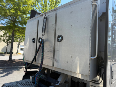 Kennworth T680 sleeper cab side panel with metal doors and latching handles; electrical harnesses and hoses hanging; part of truck body visible