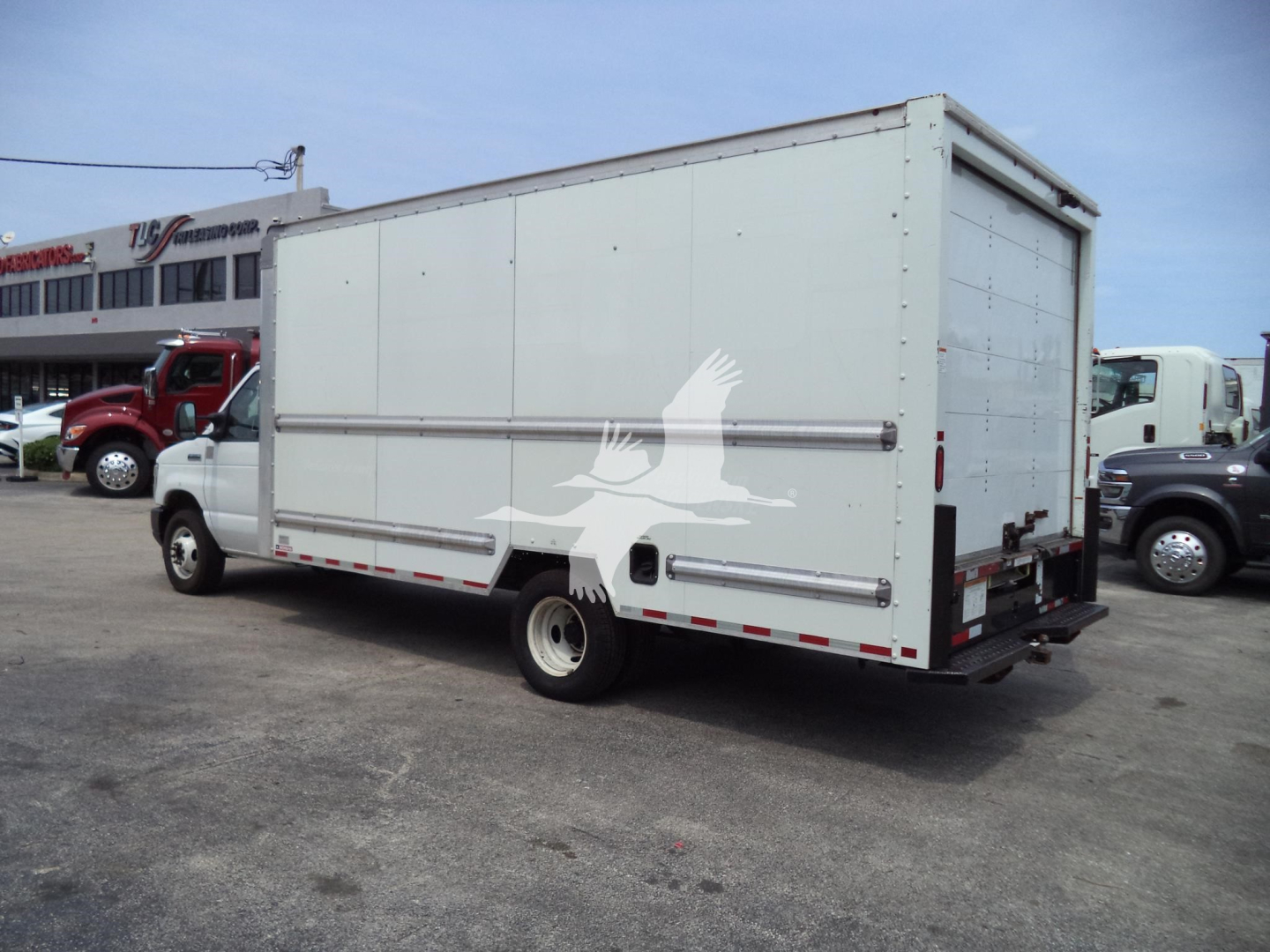 Ford E350 box truck rear three-quarter view with white cargo box, fuel tank, and dual rear doors; some text on distant building; stock/lot vehicles in background.