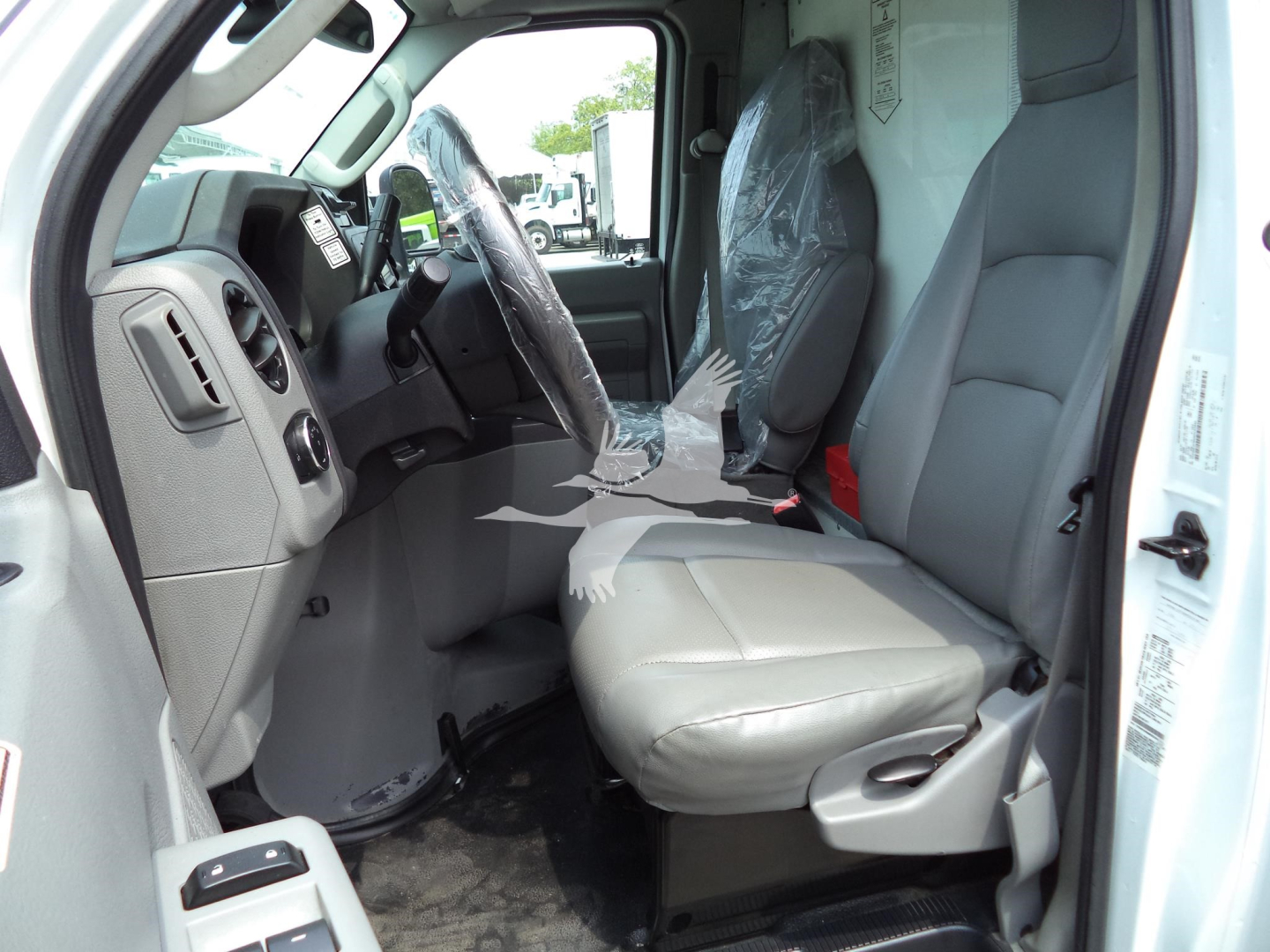Ford E-Series cargo van interior showing front driver seat with plastic cover and steering wheel, gray dashboard, and light gray seating; right-side open passenger door.